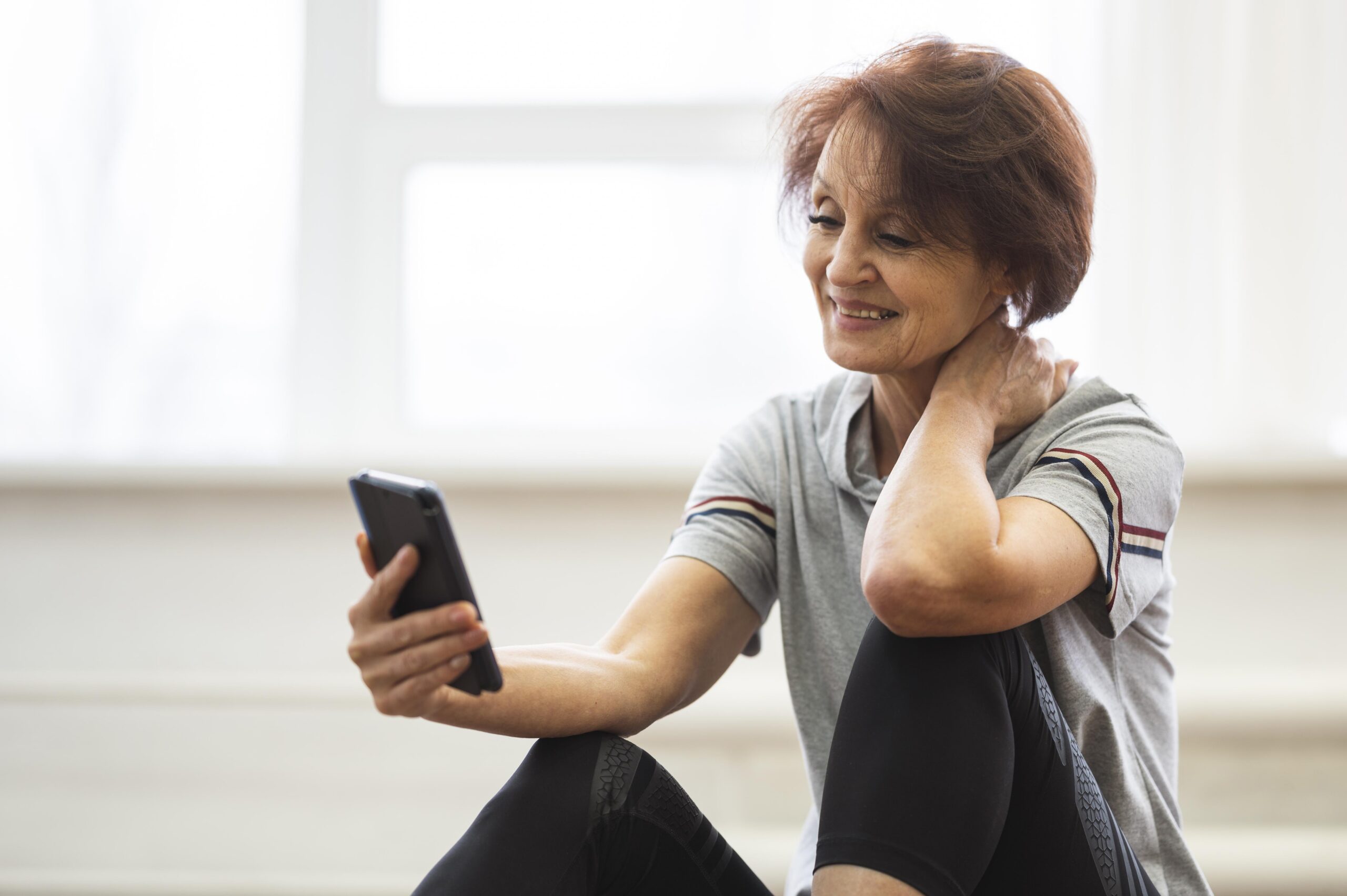 Middle‑aged woman sitting on floor using smartphone, touching her neck as if in pain, concept of checking health or arthritis app after exercise.